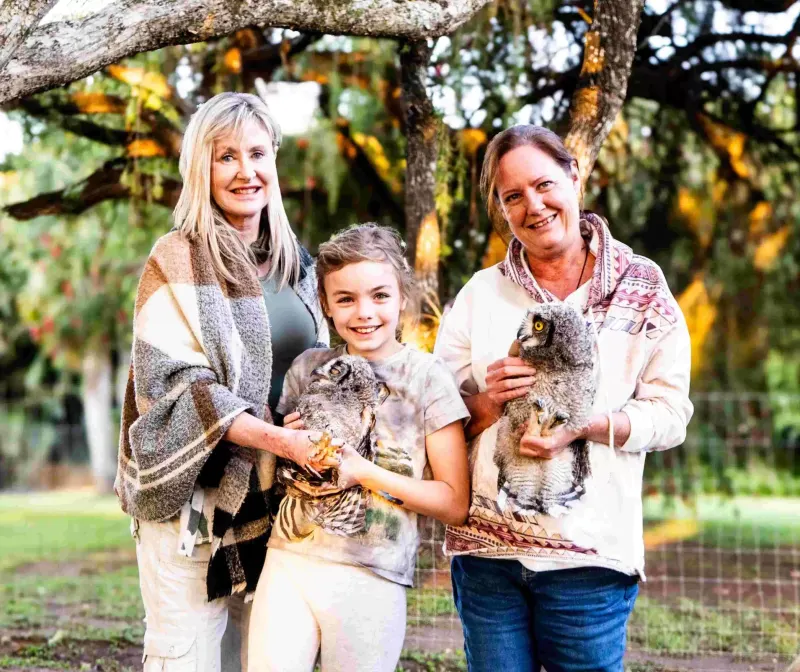 Two women and a girl smile while holding young owls outdoors beneath a large tree